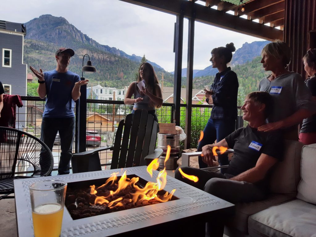 A group of climbers in casual clothes are sitting on a bar patio around a fireplace. They are drinking beer, laughing and telling stories. City skyline and mountains are in the background.